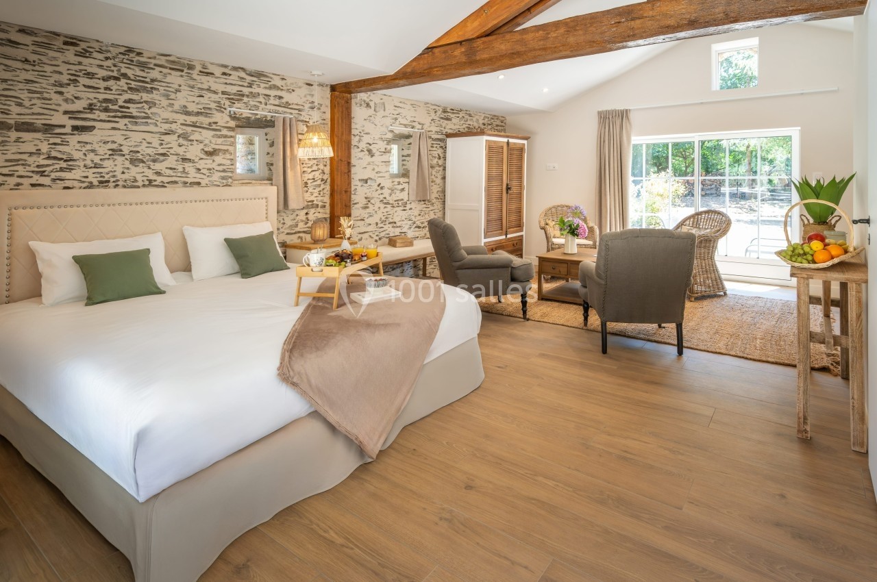 Chambre lumineuse avec lit double, fauteuils, table basse, murs en pierre apparente et grande baie vitrée donnant sur un…