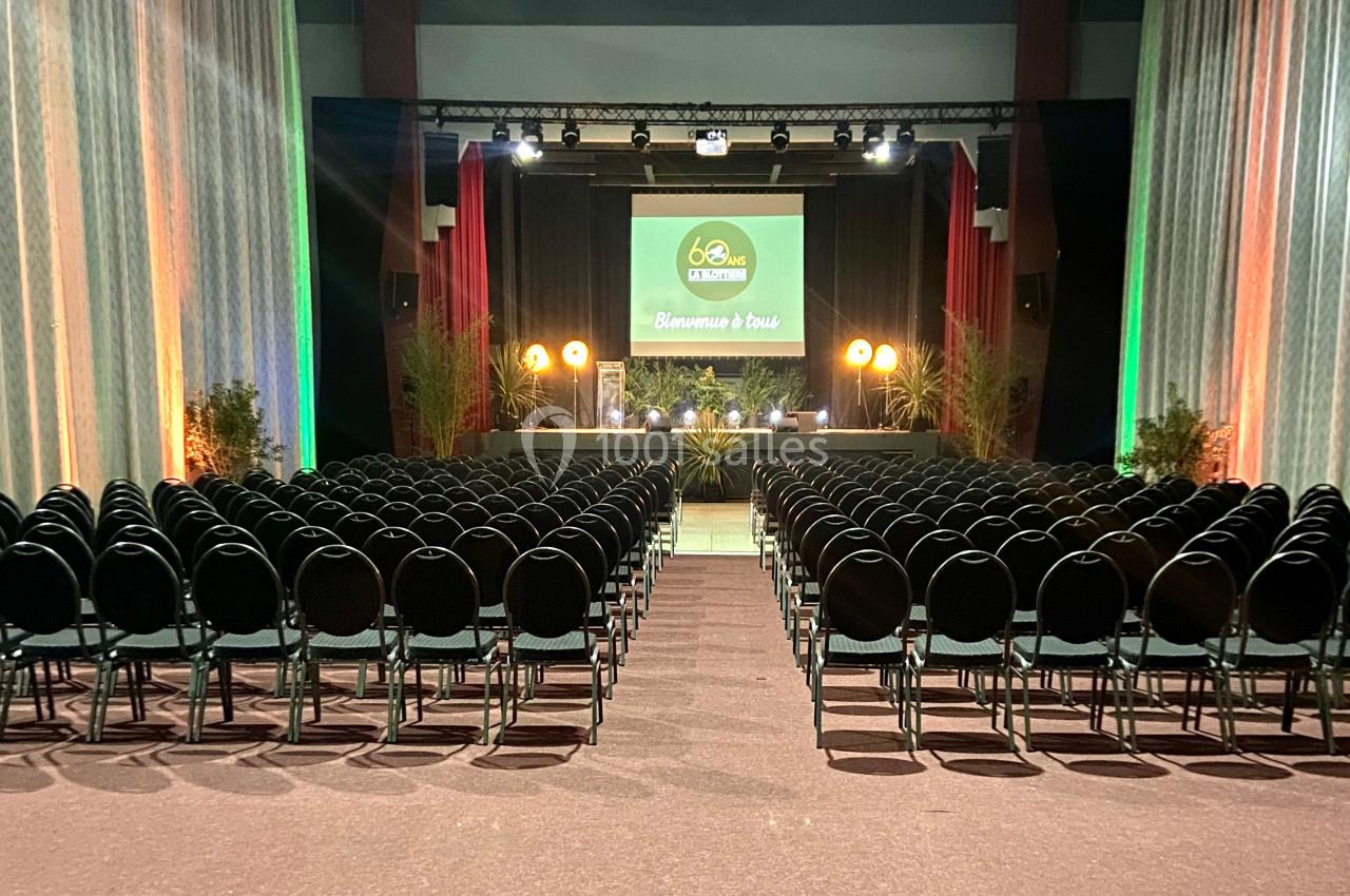Salle de conférence avec rangées de chaises noires alignées face à une scène éclairée et un écran de projection.