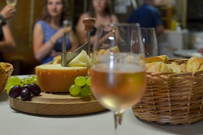 Plateau de fromage avec raisins et pain en arrière-plan, un verre de vin rosé au premier plan.