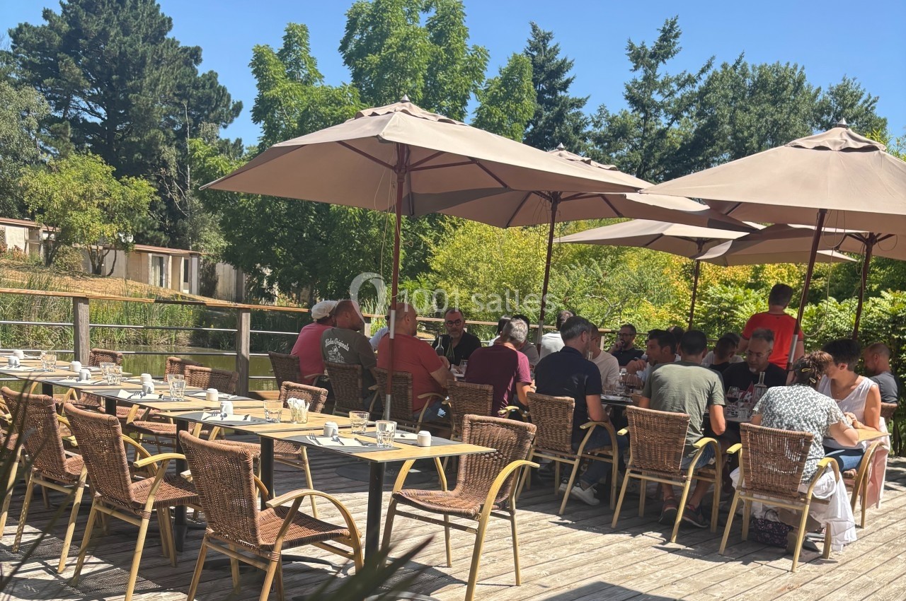 Groupe de personnes assises à des tables en terrasse ombragée, entourée de verdure par une journée ensoleillée.