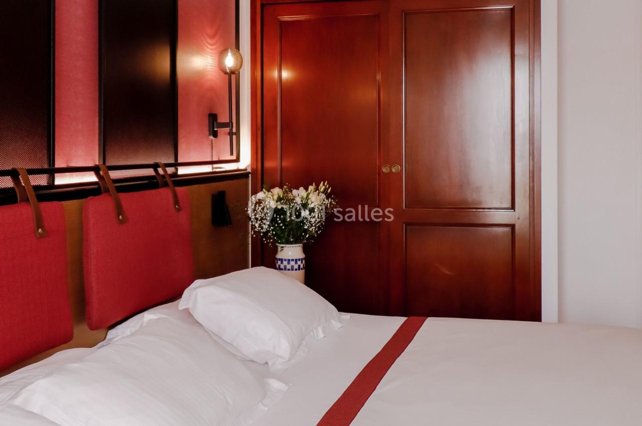 Lit avec draps blancs et tête de lit rouge, placé devant une armoire en bois et un vase de fleurs sur le côté.