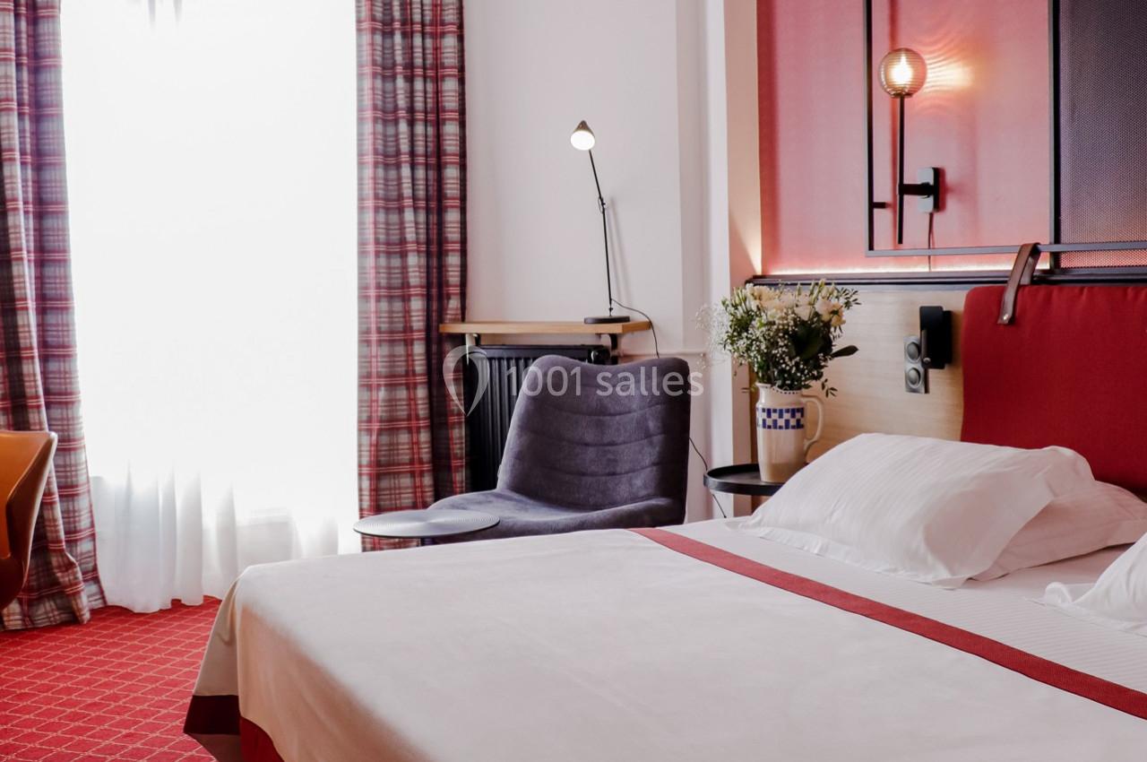 Chambre d'hôtel lumineuse avec lit double, fauteuil, lampe de lecture et rideaux blancs laissant passer la lumière.