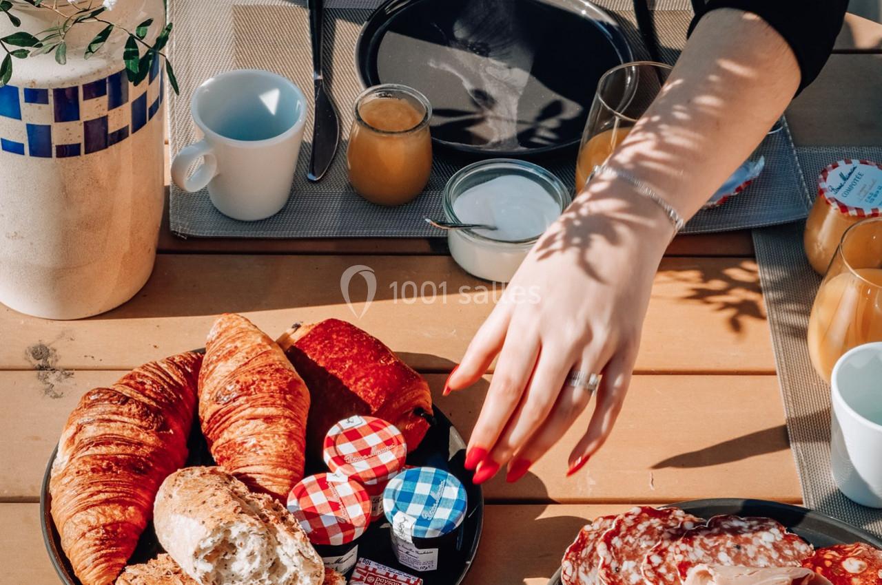 Petit-déjeuner en extérieur avec viennoiseries, confitures, charcuterie et boissons sur une table en bois.