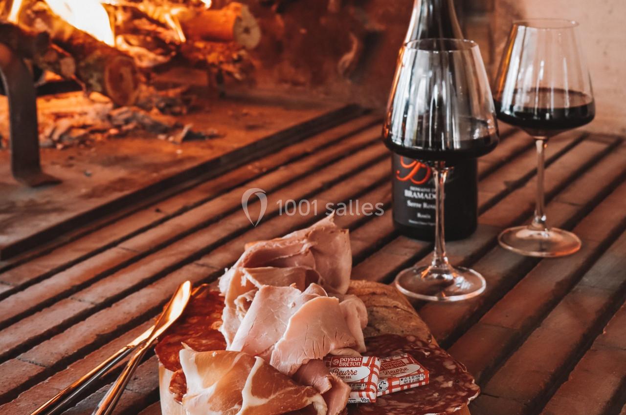 Planche de charcuterie avec jambon et saucisson, deux verres de vin rouge et une cheminée allumée en arrière-plan.