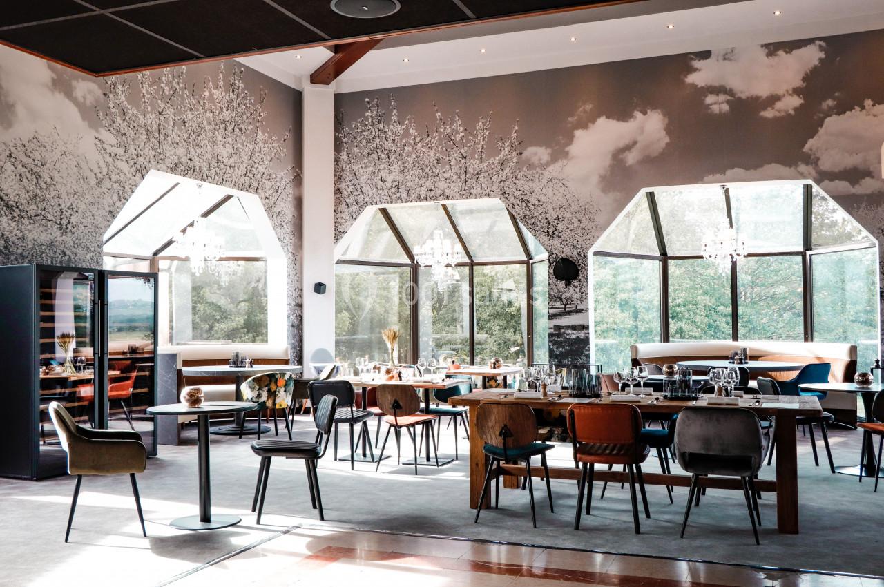 Salle de restaurant lumineuse avec de grandes baies vitrées, des tables dressées et un décor mural en noir et blanc.