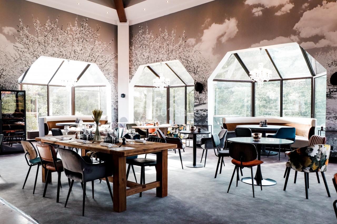Salle de restaurant lumineuse avec grandes baies vitrées, tables en bois et chaises variées dans un décor moderne.