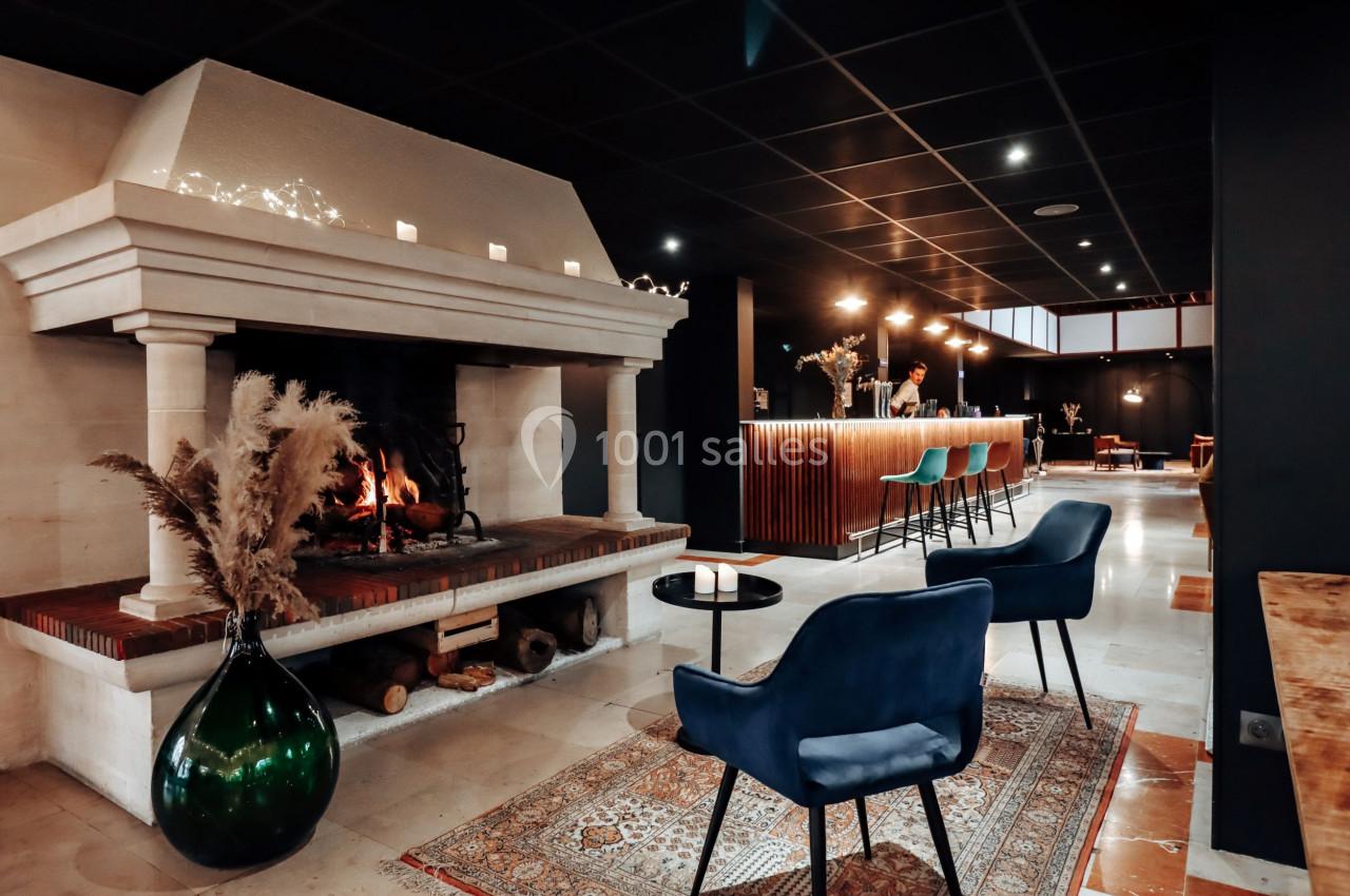 Cheminée allumée dans un espace moderne avec fauteuils bleus, tapis, et un bar éclairé en arrière-plan.