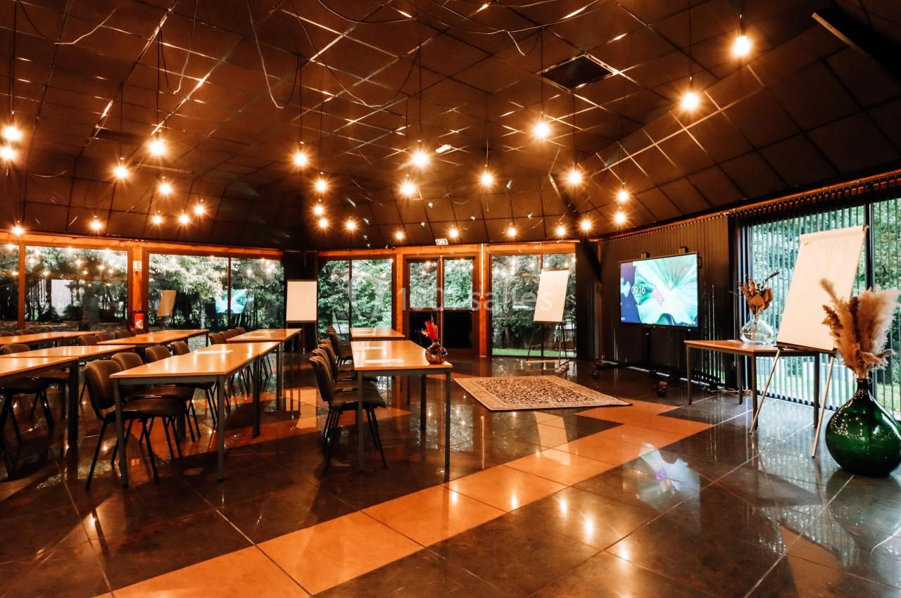 Salle de réunion spacieuse avec tables en bois, chaises, écrans, tableaux blancs et éclairage chaleureux.