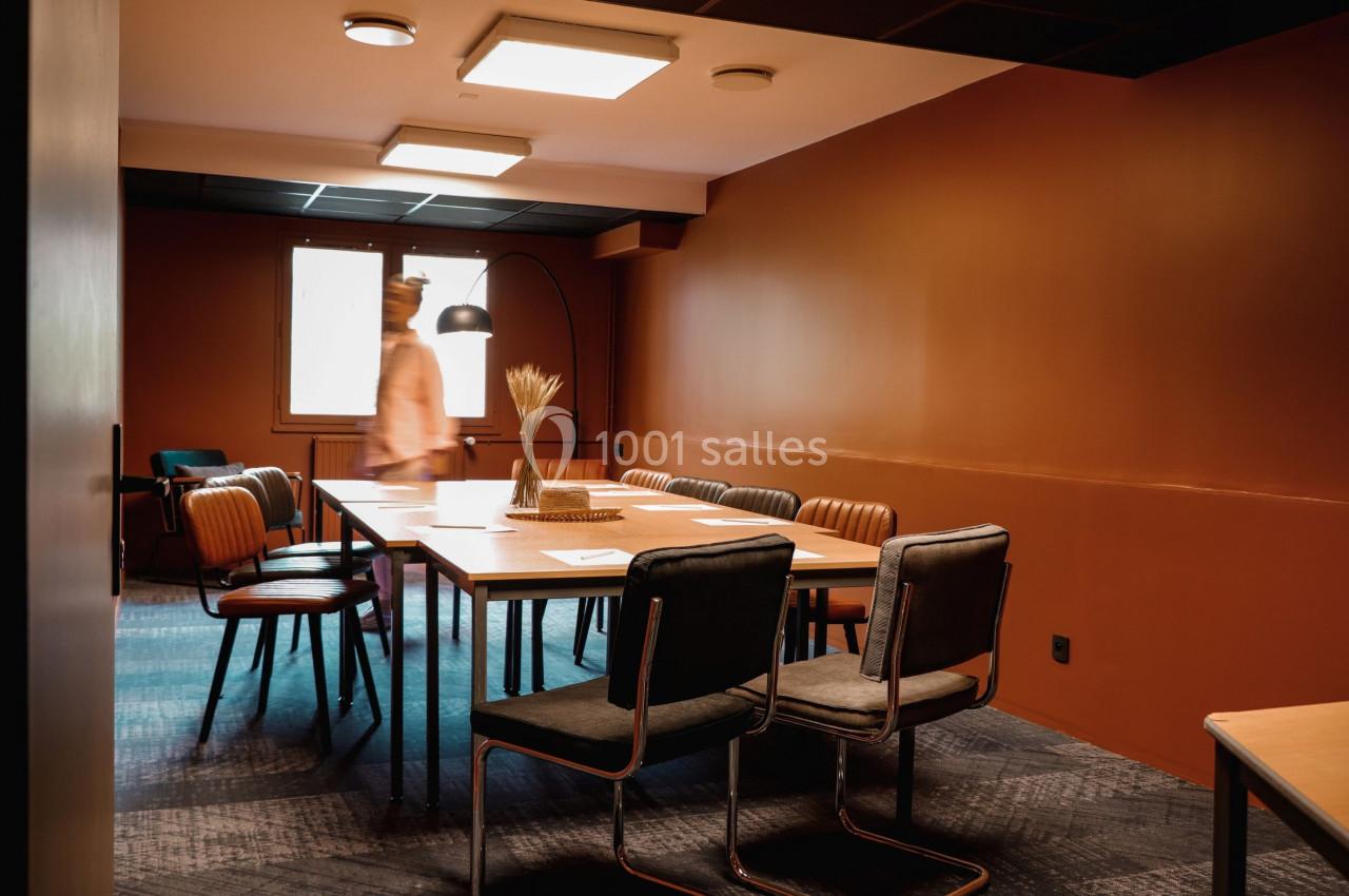 Salle de réunion avec murs orange, table en bois, chaises variées et une personne floue en mouvement.