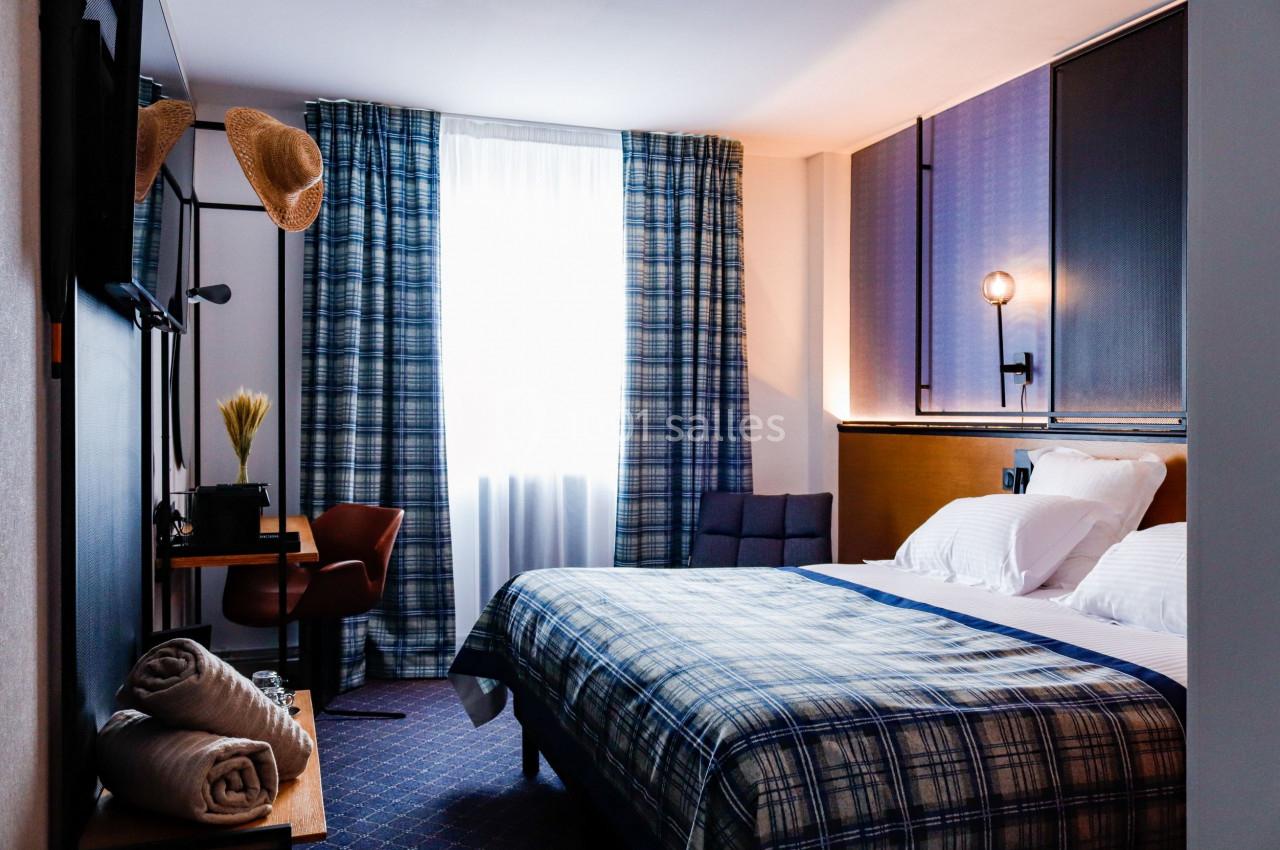 Chambre d'hôtel lumineuse avec lit double, bureau, chaise, rideaux à carreaux et décoration sobre.