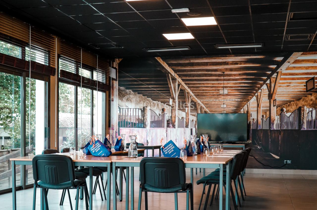 Salle lumineuse avec de grandes baies vitrées, des chaises et des tables alignées, décorée d'une fresque représentant une…