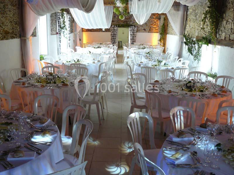 Salle de réception décorée avec des tables rondes dressées, nappes blanches, chaises en plastique et éclairage tamisé.