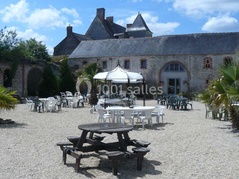 Cour extérieure avec tables, chaises et tonnelle, entourée de bâtiments en pierre sous un ciel partiellement nuageux.