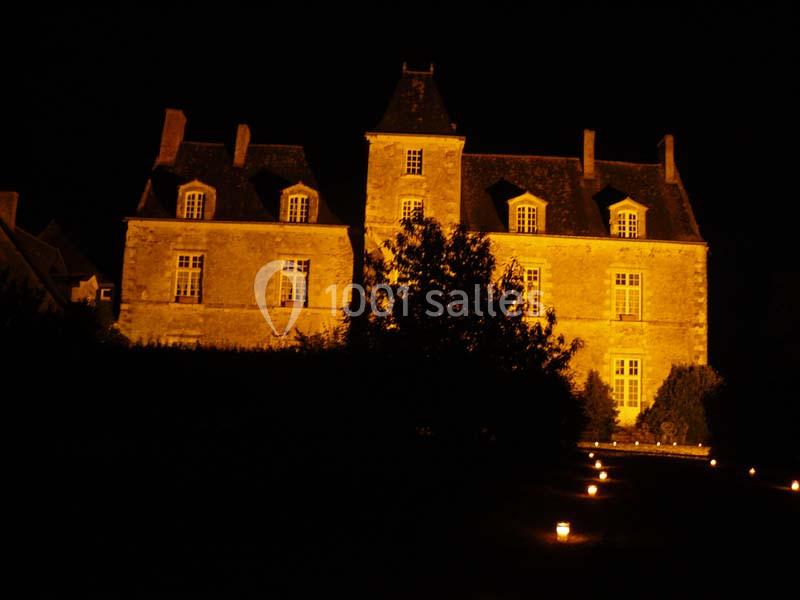 Façade d'un manoir éclairé par des lumières chaudes, entouré de végétation et de bougies au sol, de nuit.