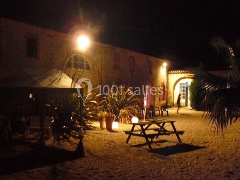 Cour éclairée d'une bâtisse en pierre la nuit, avec table de pique-nique, plantes et lumière tamisée.