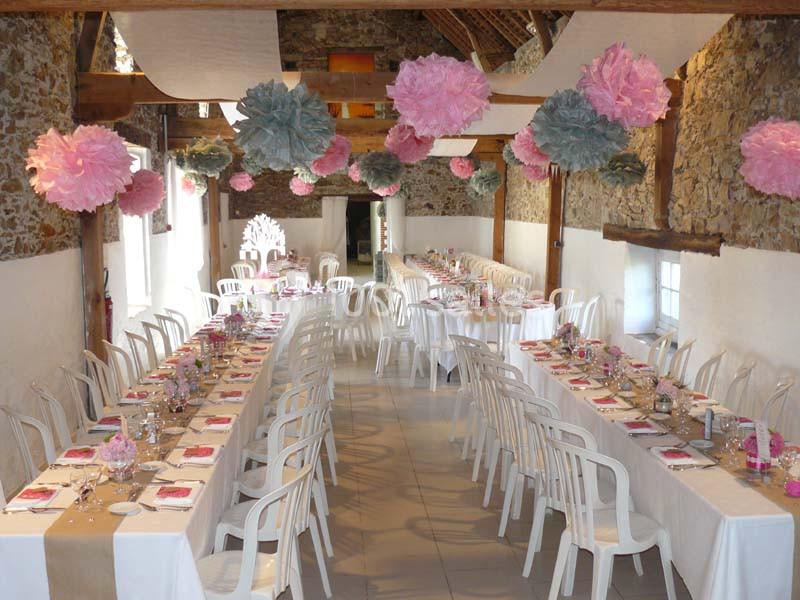 Salle décorée pour un événement avec tables alignées, nappes blanches, chaises en plastique et suspensions en papier coloré.