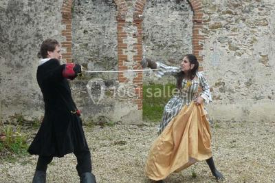 Deux personnes en costume historique s'affrontent à l'épée dans une cour devant un mur en pierre avec des arches.
