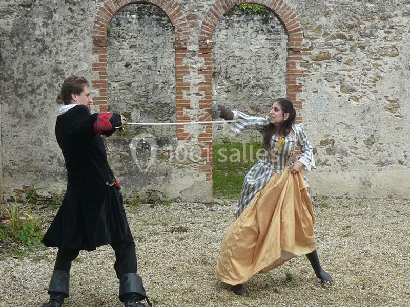 Deux personnes en costume historique s'affrontent à l'épée dans une cour devant un mur en pierre avec des arches.