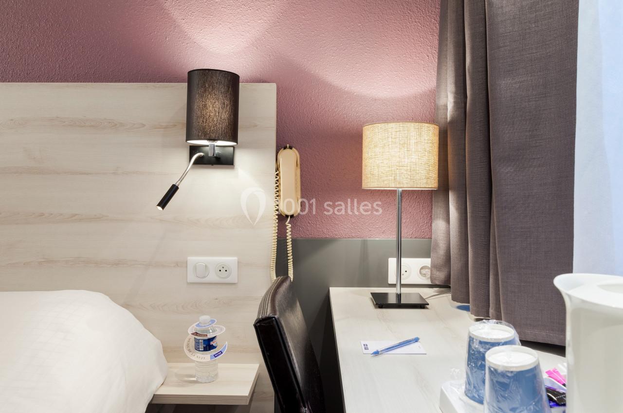 Lampe de chevet et lampe de table allumées sur un bureau près d'un lit, avec téléphone, gobelets et rideaux.