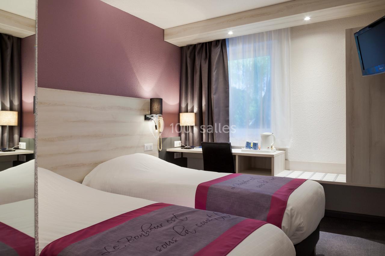 Chambre d'hôtel avec deux lits simples, bureau, télévision murale, miroir et fenêtre avec rideaux.