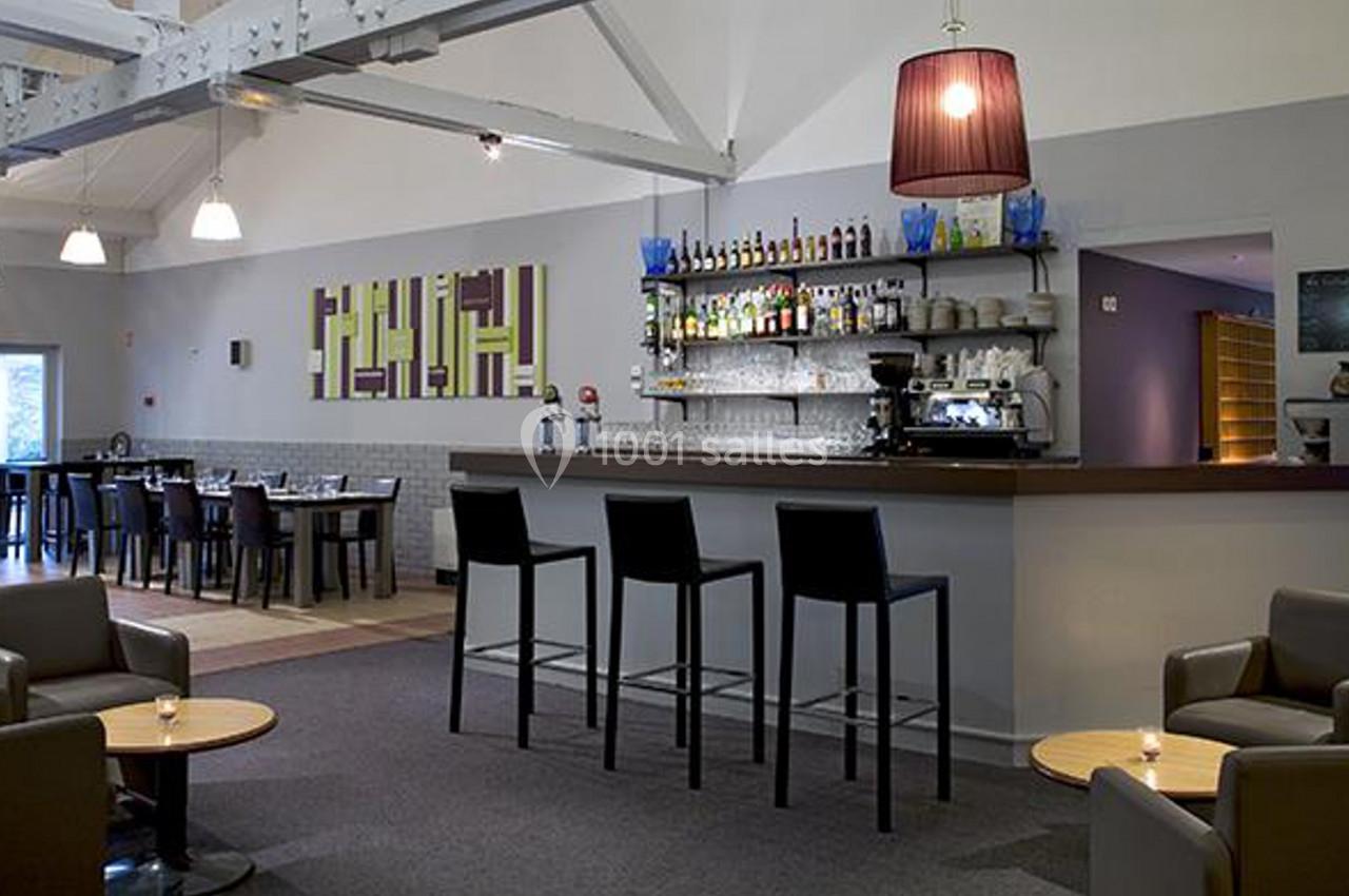 Bar moderne avec comptoir en bois, tabourets hauts, étagères de bouteilles, et coin salon avec fauteuils et tables basses.