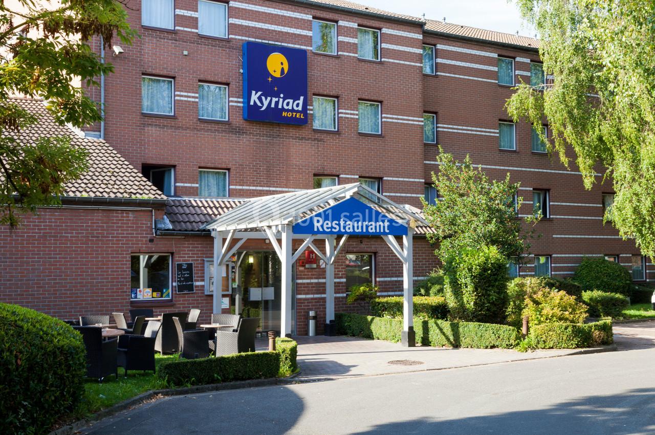 Façade d'un hôtel en briques rouges avec un panneau ’Kyriad Hotel’ et une entrée de restaurant entourée de verdure.