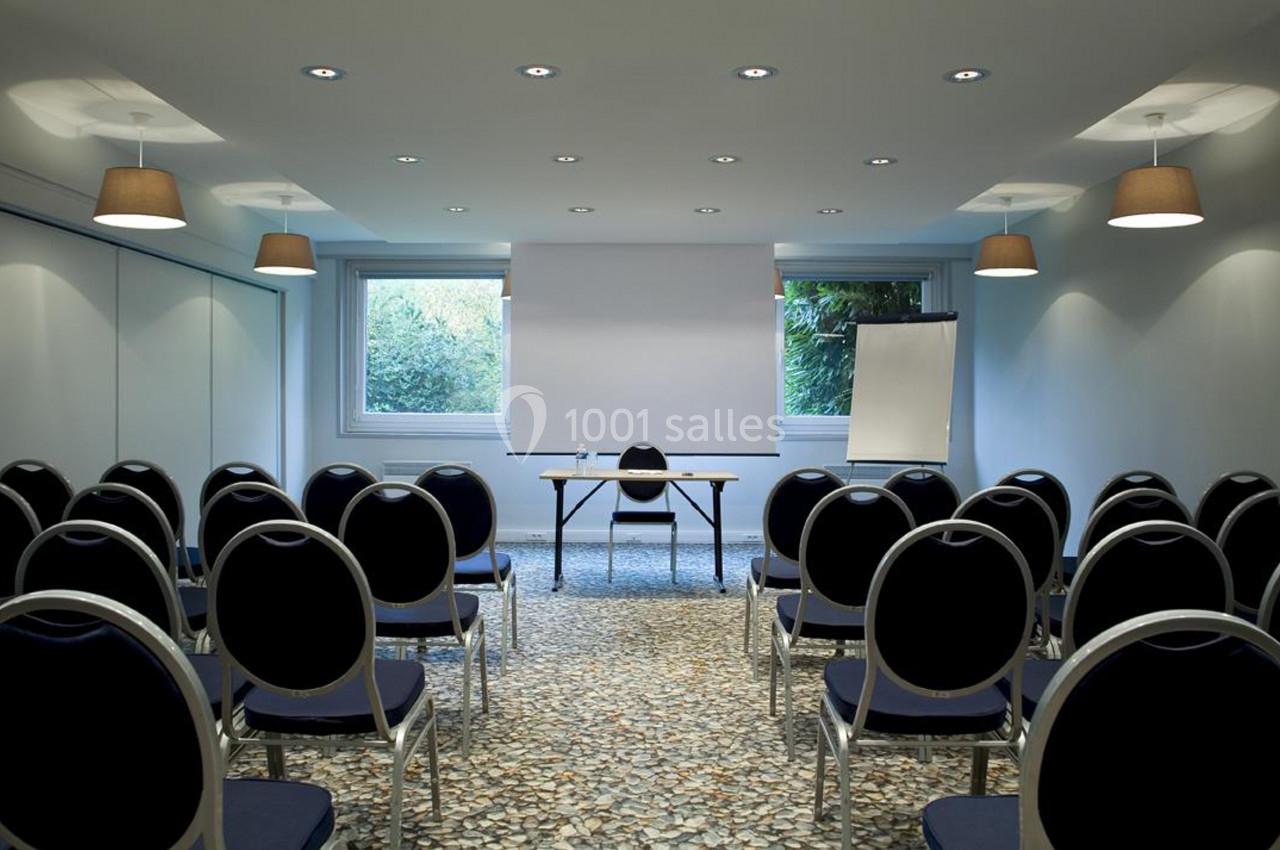 Salle de réunion lumineuse avec des rangées de chaises noires, une table, un écran de projection et un tableau blanc.