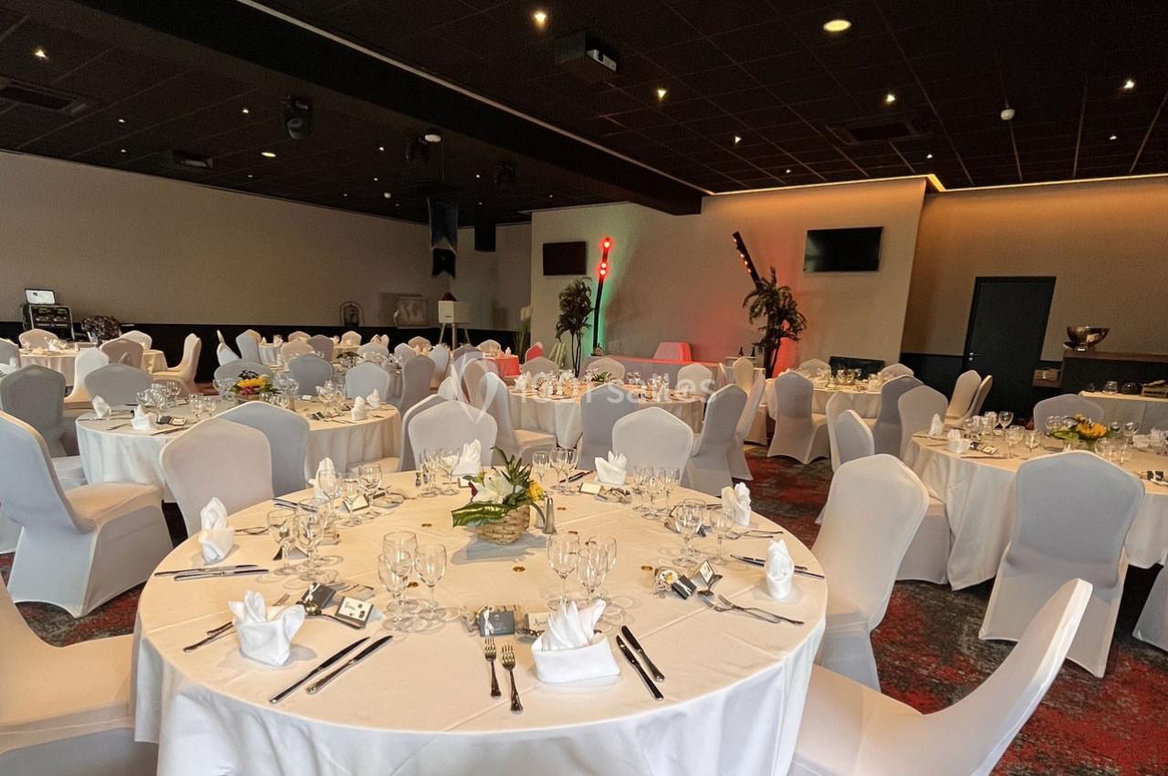 Salle de réception avec tables rondes dressées, nappes blanches, chaises assorties et décorations florales.