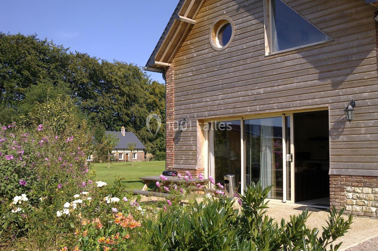 Maison en bois avec de grandes baies vitrées, entourée d'un jardin fleuri et d'arbres en arrière-plan.