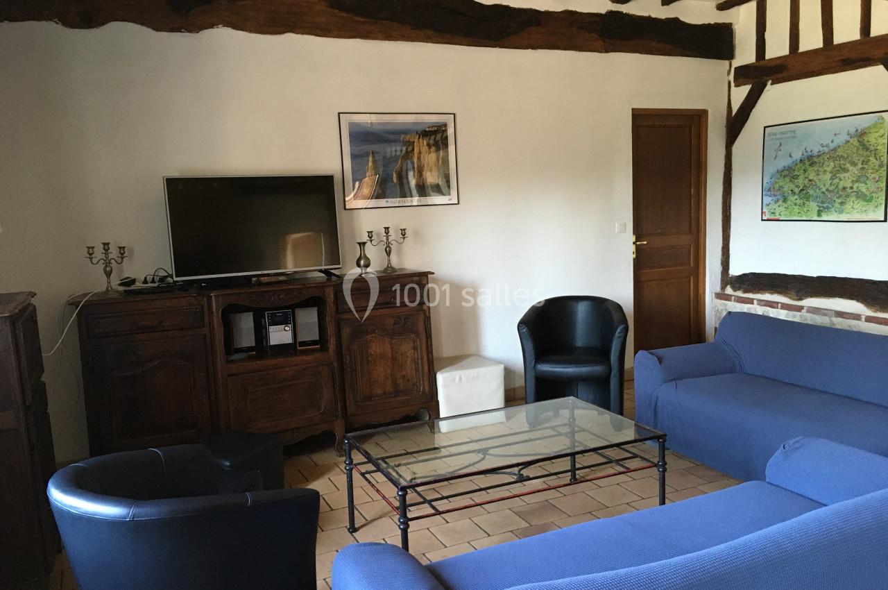 Salon avec poutres apparentes, canapé bleu, fauteuils noirs, table basse en verre et meuble TV en bois.