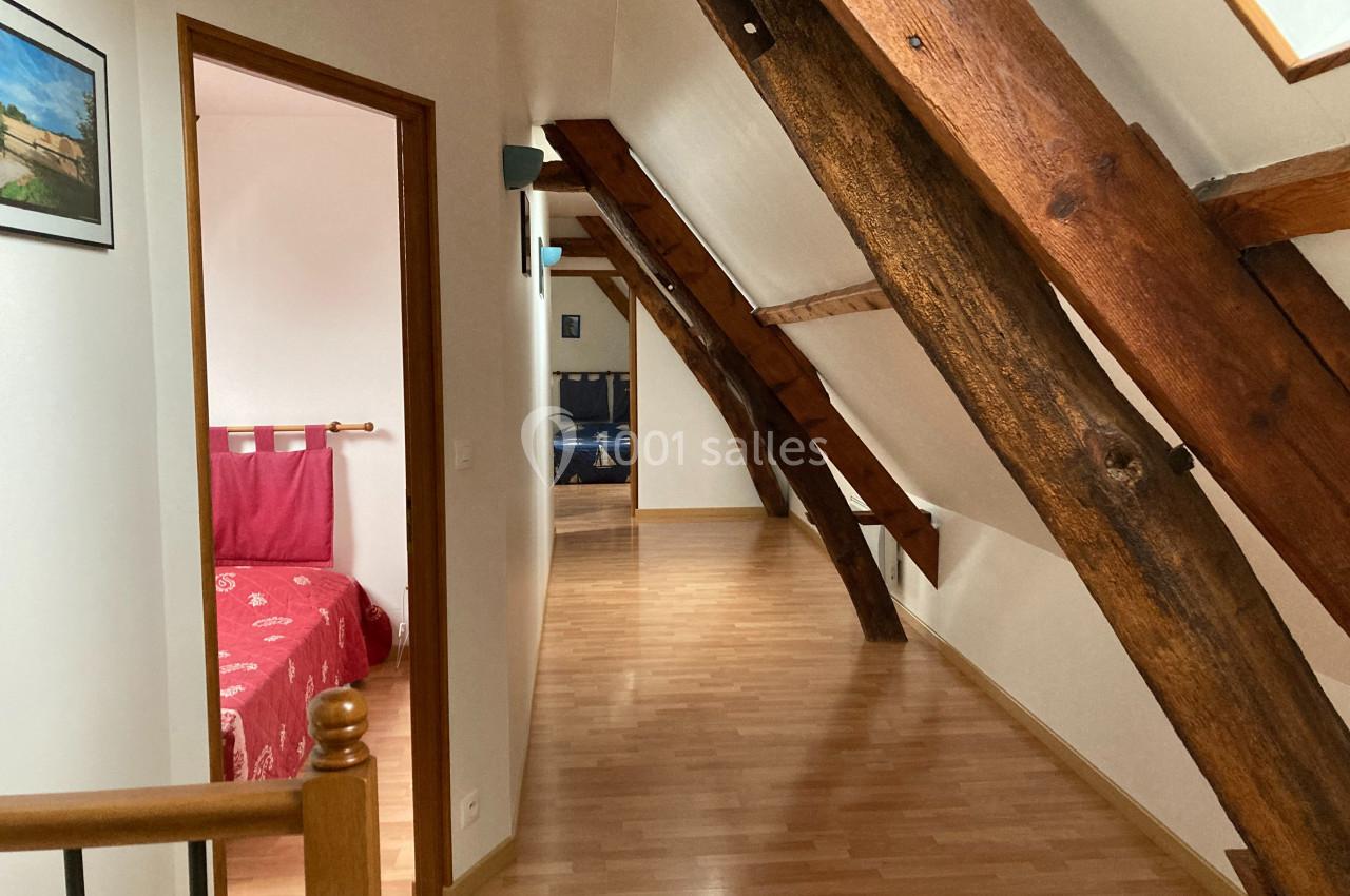 Vue d'un couloir mansardé avec poutres apparentes, menant à deux chambres aux lits visibles en arrière-plan.