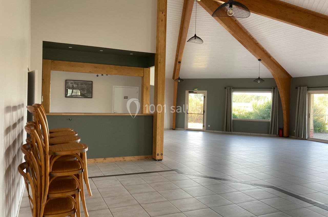 Salle spacieuse avec charpente en bois, sol carrelé, chaises empilées à gauche et grandes fenêtres laissant entrer la…