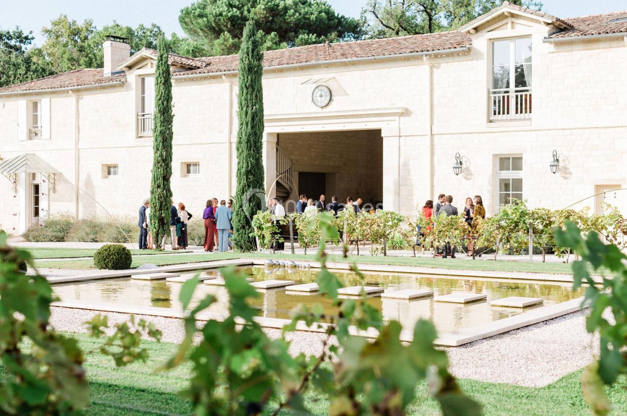 Extérieur Château Gassies Des personnes rassemblées devant un bâtiment en pierre clair, entouré de jardins avec un bassin et des vignes au premier…