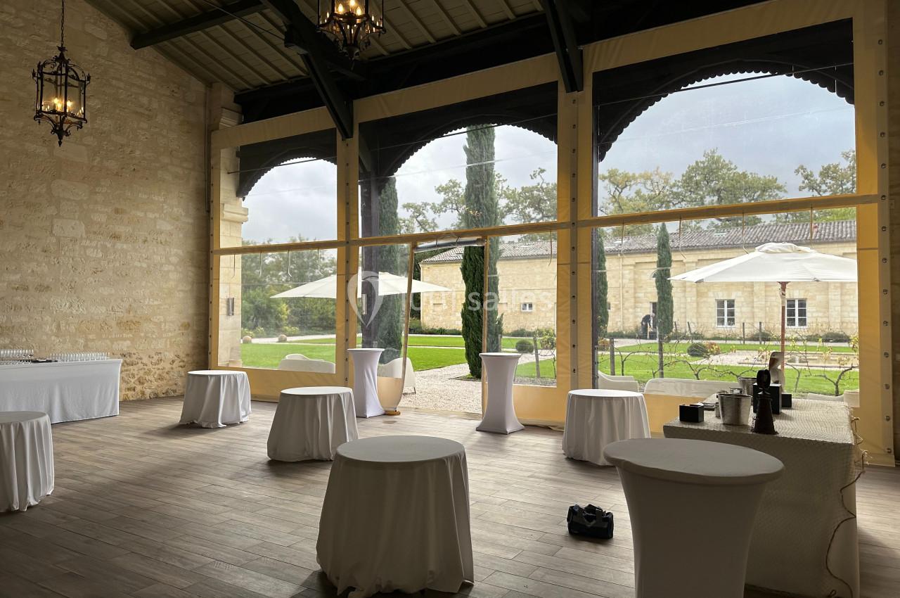 Préau Orangerie Salle lumineuse avec tables hautes drapées de blanc, grandes baies vitrées donnant sur un jardin avec parasols.