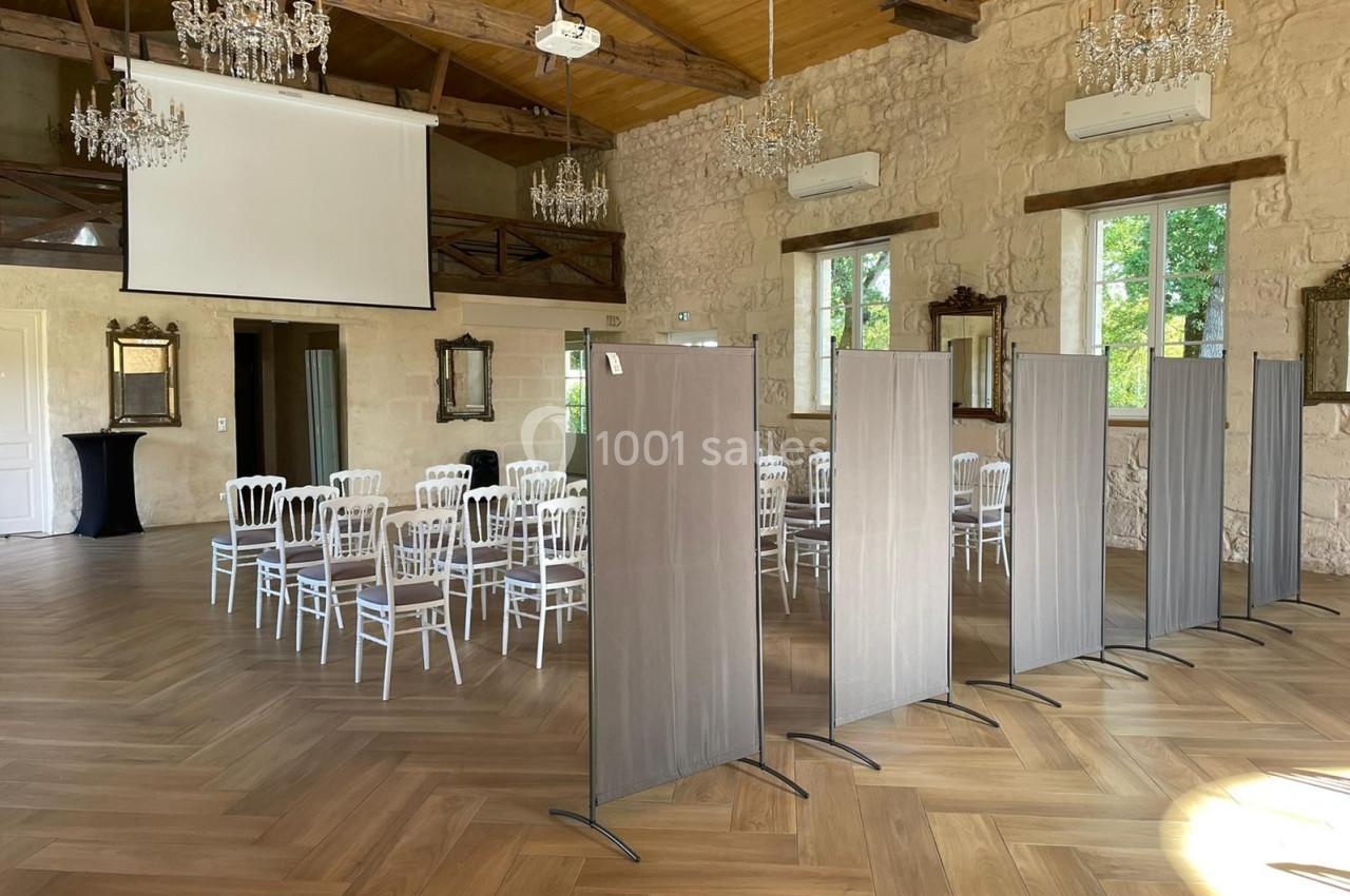 Salle de Réception - 190m² Salle lumineuse avec parquet, chaises blanches disposées en rangées, écrans de séparation et projecteur au plafond.