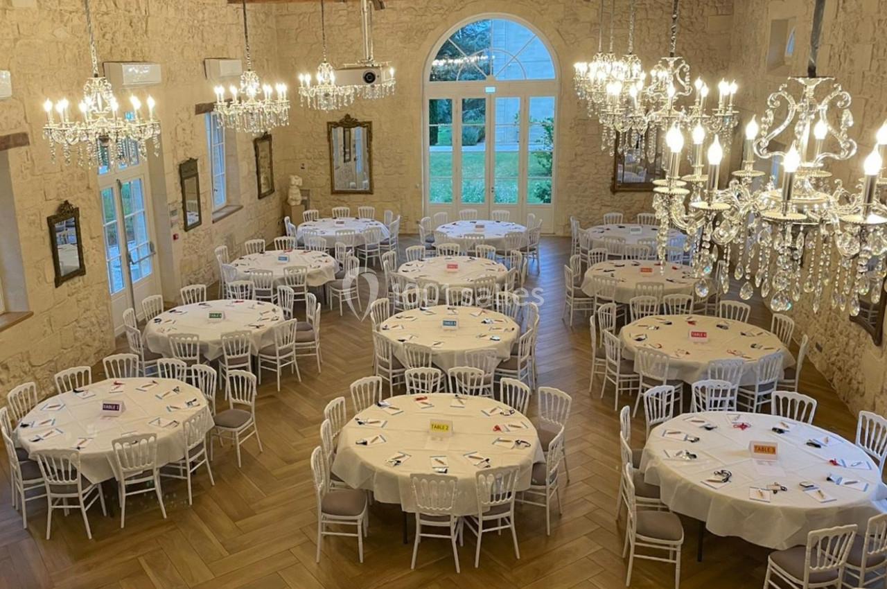 Salle de Réception - 190m² Salle de réception avec tables rondes dressées, chaises blanches, lustres en cristal et grandes fenêtres donnant sur un…