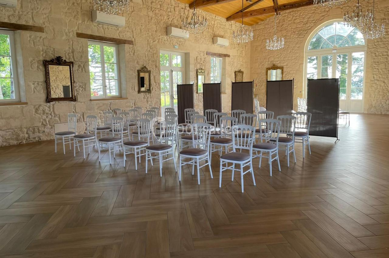 Salle de Réception - 190m² Salle lumineuse avec parquet en bois, chaises blanches disposées en cercle et paravents noirs devant des fenêtres.