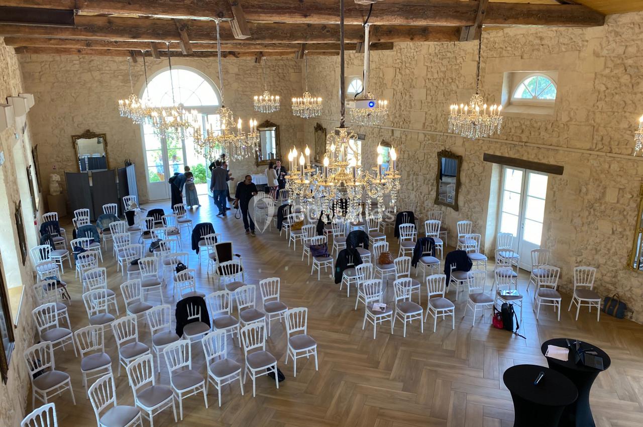 Salle de Réception - 190m² Salle en pierre avec des chaises blanches disposées pour un événement, éclairée par des lustres et de grandes fenêtres.
