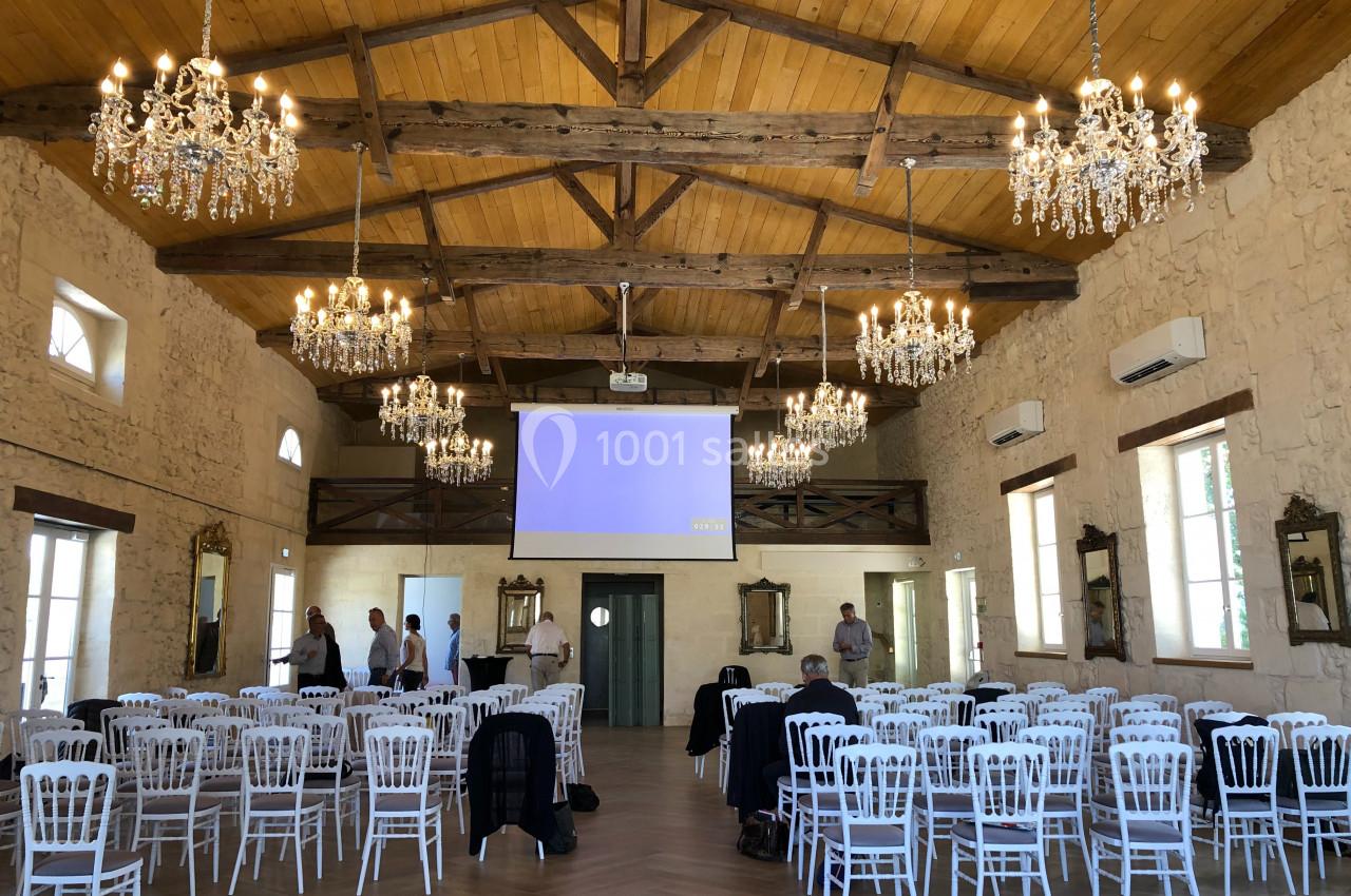 Salle de Réception - 190m² Salle de réception avec poutres apparentes, lustres, chaises blanches alignées et écran de projection allumé.