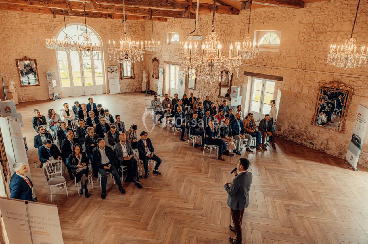 Salle de Réception - 190m² Un conférencier s'adresse à un public assis dans une salle lumineuse avec des murs en pierre et des lustres.