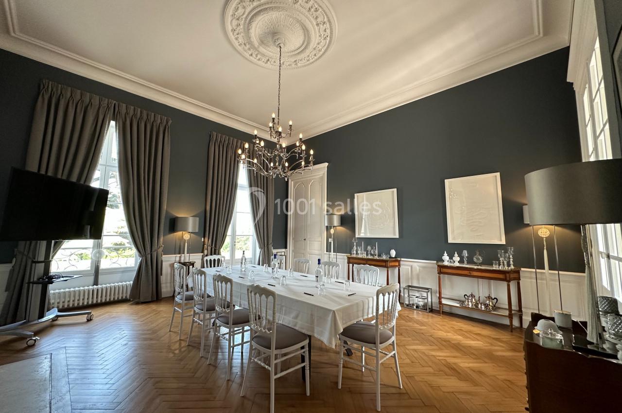 Salon Gris Privé Salle à manger élégante avec table dressée, chaises blanches, lustre central, murs bleu foncé et parquet en bois clair.