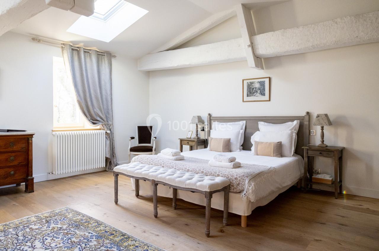 Suite Tilleul Chambre lumineuse avec lit double, poutres apparentes, parquet, tapis et fenêtre avec rideaux gris.