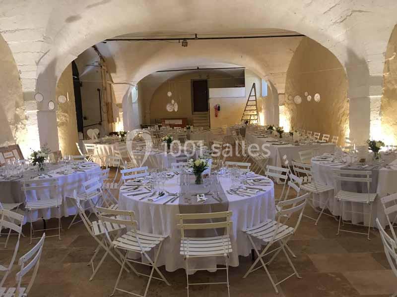 Salle voûtée décorée pour un événement avec des tables rondes dressées, des chaises blanches et des éclairages chaleureux.