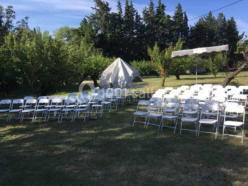 Chaises blanches disposées en rangées sur une pelouse, avec une tente et des arbres en arrière-plan.