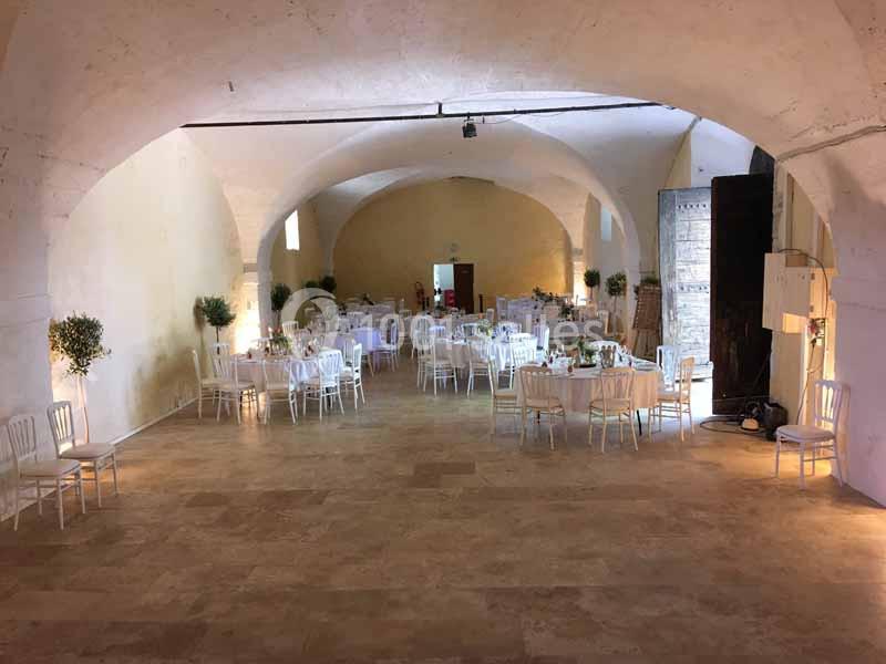 Salle voûtée décorée pour un événement, avec tables rondes dressées, chaises blanches et plantes en pot.