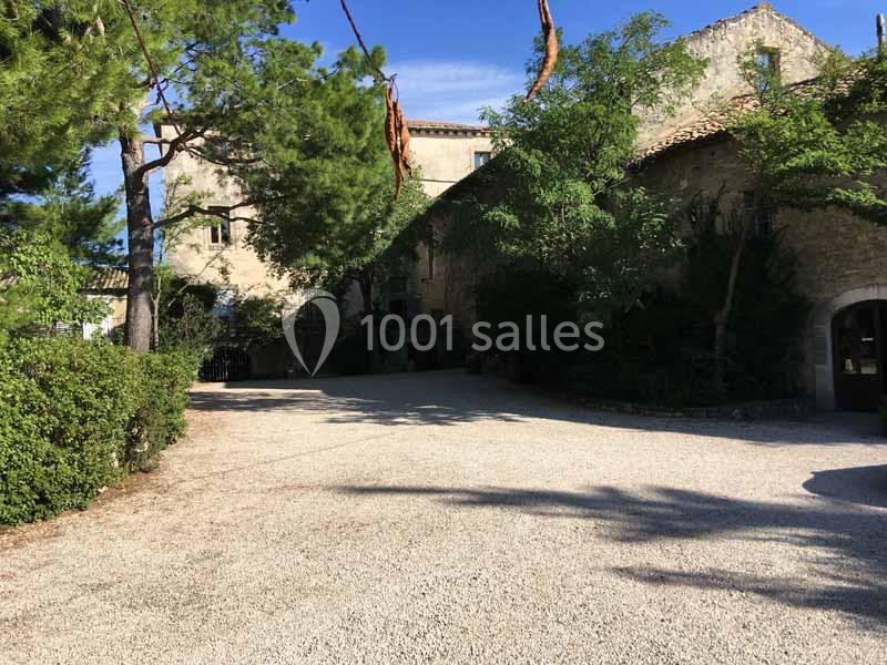 Cour en gravier bordée d'arbres et d'un bâtiment en pierre avec des volets, sous un ciel bleu.