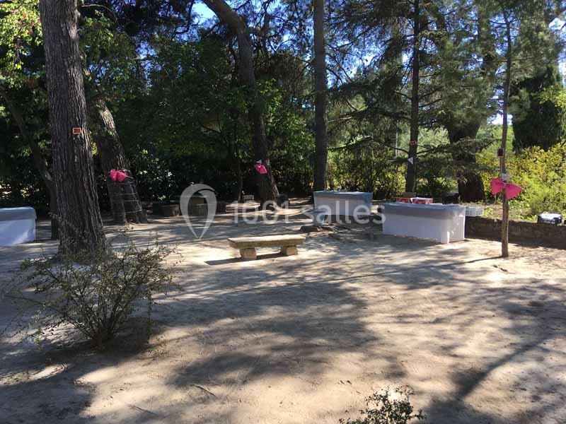 Espace extérieur ombragé avec banc en pierre, tables décorées de nappes blanches et arbres environnants.