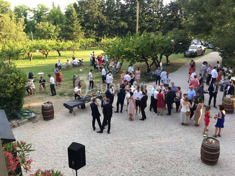 Personnes rassemblées en extérieur dans une cour gravillonnée entourée d'arbres, lors d'un événement convivial.