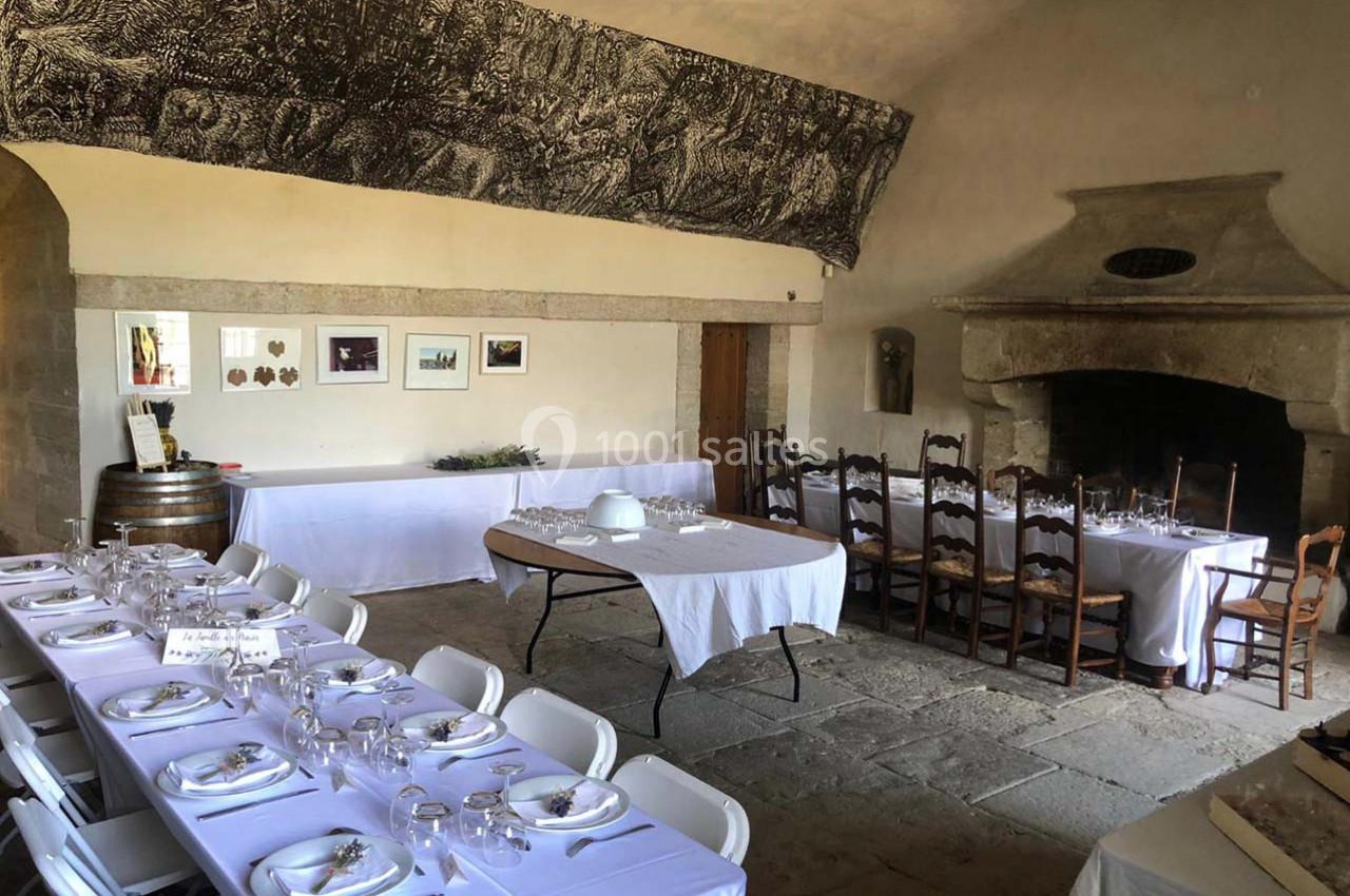 Salle en pierre avec tables dressées pour un repas, nappes blanches, chaises pliantes et cheminée ancienne.