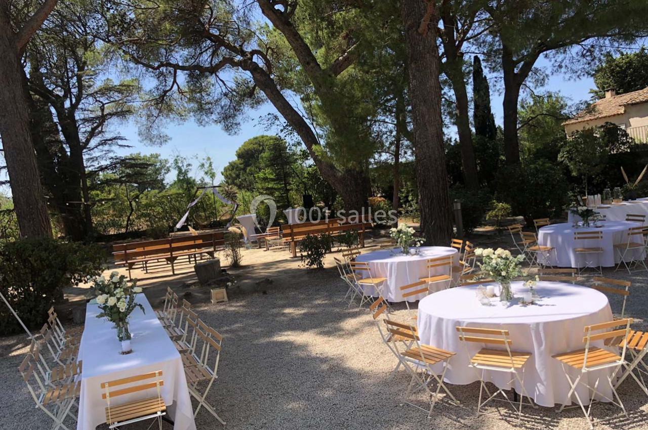 Tables rondes décorées de fleurs blanches et chaises en bois disposées en extérieur sous des arbres dans un cadre ensoleillé.